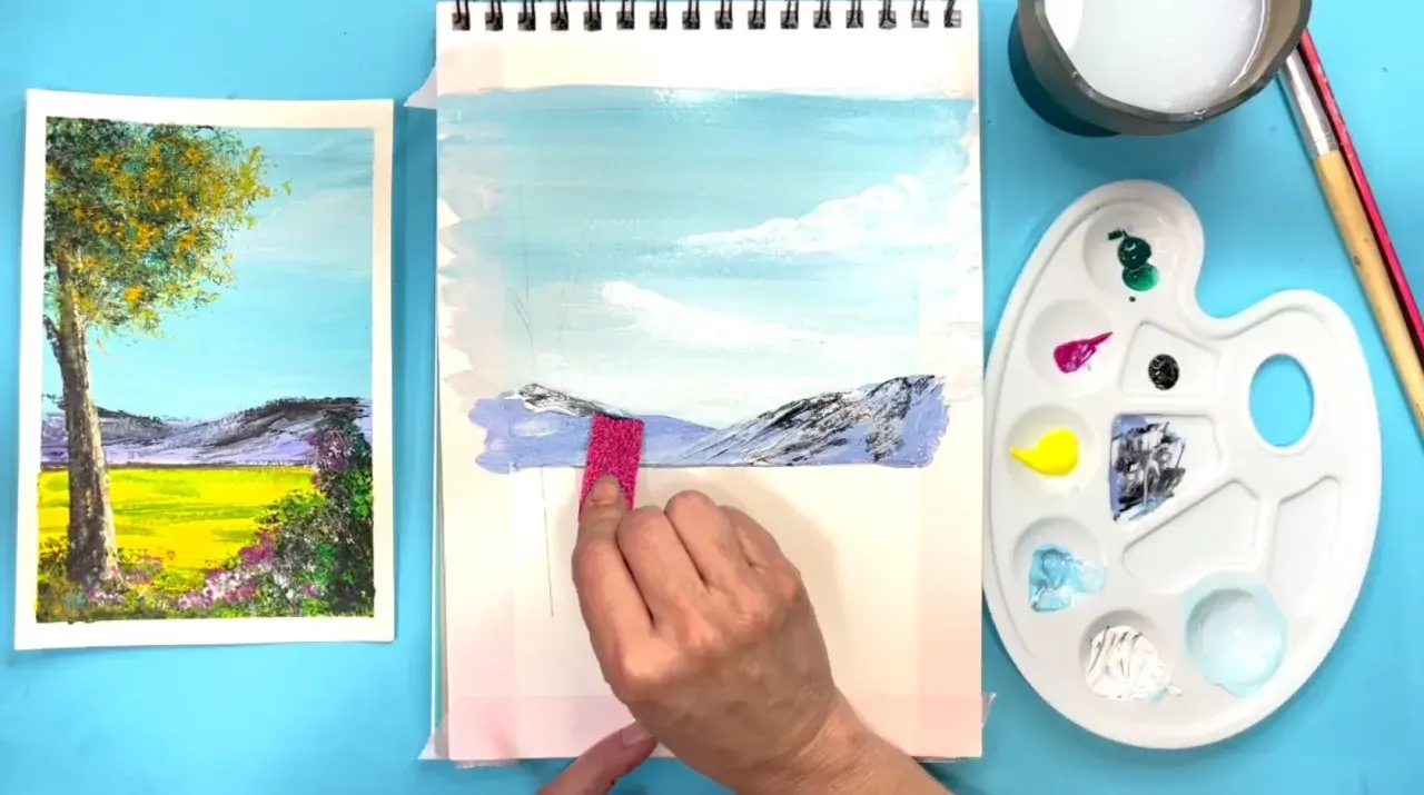 Creando dibujos de paisajes fáciles. Una mano aplica pintura a un lienzo con montañas y cielo azul.