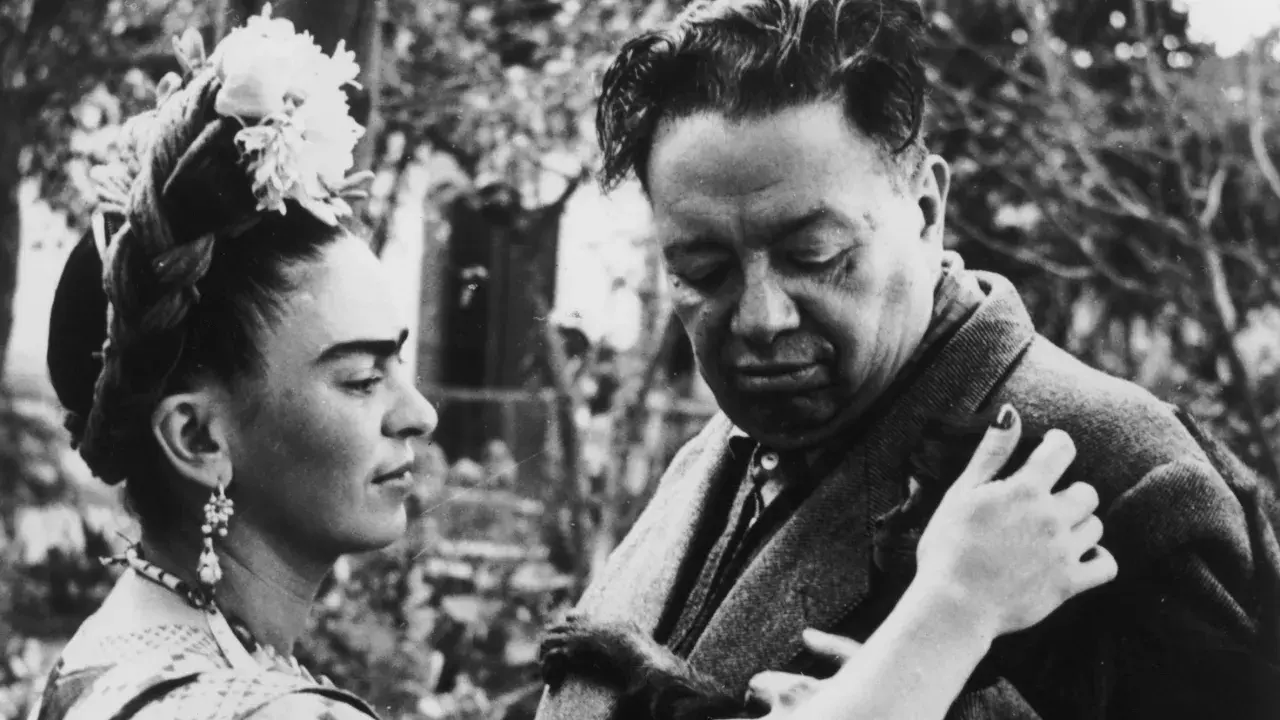 Frida Kahlo y Diego Rivera, una pareja icónica, comparten un momento íntimo. Frida, con flores en el cabello, mira a Diego, quien parece reflexivo.