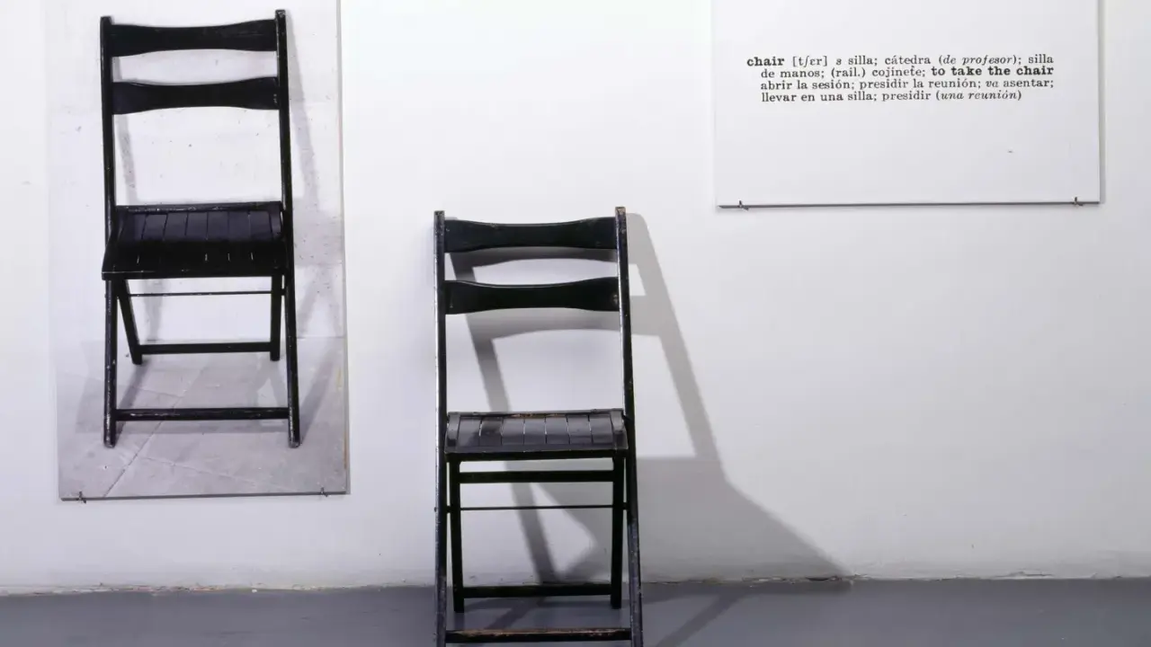 Una y tres sillas. Dos sillas negras plegables, una colgada en la pared y otra de pie, junto a un cartel con la definición de "chair".