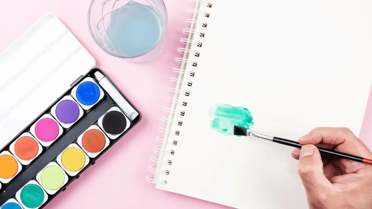 Una mano aplica pintura en acuarela verde en un cuaderno. A su lado, una caja de acuarelas y un vaso con agua azul.
