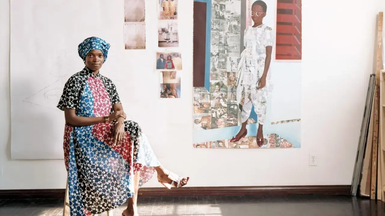 Njideka Akunyili Crosby, sentada en un taburete, luce un vestido vibrante y un turbante. Detrás, obras de arte y fotos familiares adornan la pared.