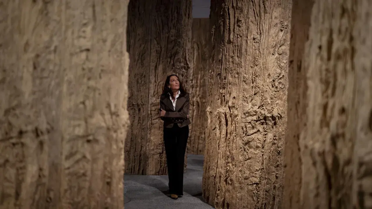 Cristina Iglesias, rodeada de imponentes estructuras de madera que evocan un bosque, contempla la obra.