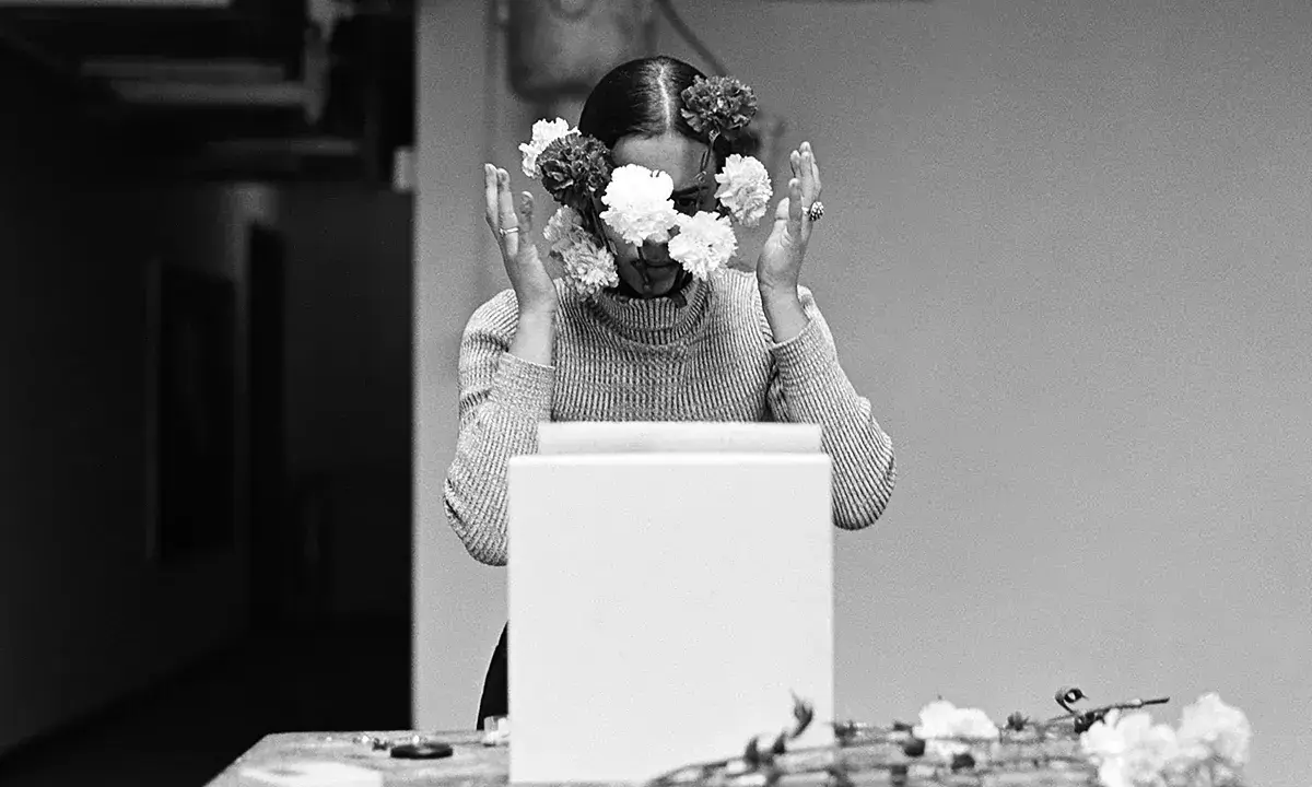 Una mujer con flores en el rostro, evocando las **obras de Ana Mendieta**. Sus manos se alzan, rodeando su cabeza adornada con pétalos blancos y oscuros.