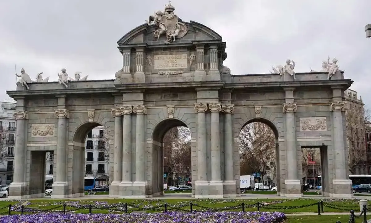 La majestuosa Puerta de Alcalá se alza imponente, con sus arcos y esculturas, rodeada de un jardín de flores moradas.