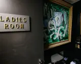 Señal de "Ladies Room" dorada sobre pared oscura, junto a pintura abstracta y retrete.