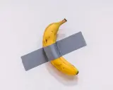 Una obra de arte platano, un plátano amarillo pegado a una pared blanca con cinta adhesiva gris.