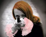 Mujer pelirroja apunta con una pistola, con toques rojos en su ropa, evocando las audaces obras de arte de Niki de Saint Phalle.
