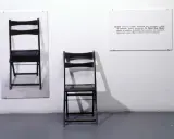 Una y tres sillas. Dos sillas negras plegables, una colgada en la pared y otra de pie, junto a un cartel con la definición de "chair".