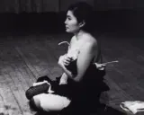 Yoko Ono, en el escenario, sentada en el suelo, con ropa desprendida. Una escena icónica de Yoko Ono Cut Piece.