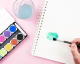 Una mano aplica pintura en acuarela verde en un cuaderno. A su lado, una caja de acuarelas y un vaso con agua azul.