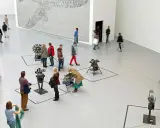 Gente pasea por una galería de arte, observando instalaciones mecánicas y un gran mural. Un día en las ferias de arte.