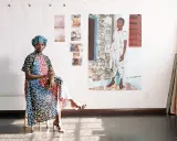 Njideka Akunyili Crosby, sentada en un taburete, luce un vestido vibrante y un turbante. Detrás, obras de arte y fotos familiares adornan la pared.