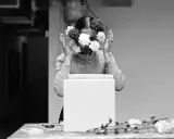 Una mujer con flores en el rostro, evocando las **obras de Ana Mendieta**. Sus manos se alzan, rodeando su cabeza adornada con pétalos blancos y oscuros.