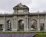 La majestuosa Puerta de Alcalá se alza imponente, con sus arcos y esculturas, rodeada de un jardín de flores moradas.