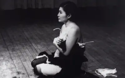 Yoko Ono, en el escenario, sentada en el suelo, con ropa desprendida. Una escena icónica de Yoko Ono Cut Piece.