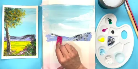 Creando dibujos de paisajes fáciles. Una mano aplica pintura a un lienzo con montañas y cielo azul.