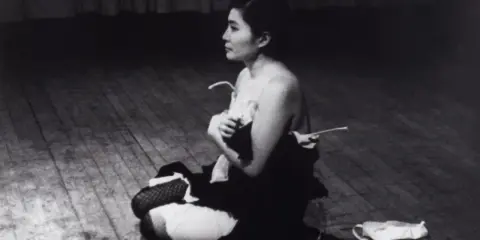 Yoko Ono, en el escenario, sentada en el suelo, con ropa desprendida. Una escena icónica de Yoko Ono Cut Piece.