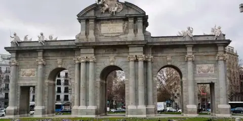 La majestuosa Puerta de Alcalá se alza imponente, con sus arcos y esculturas, rodeada de un jardín de flores moradas.