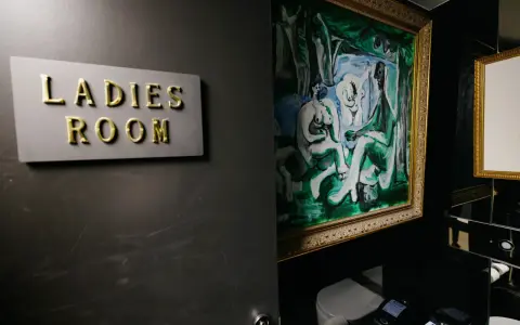 Señal de "Ladies Room" dorada sobre pared oscura, junto a pintura abstracta y retrete.