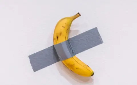 Una obra de arte platano, un plátano amarillo pegado a una pared blanca con cinta adhesiva gris.