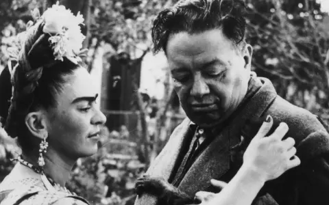Frida Kahlo y Diego Rivera, una pareja icónica, comparten un momento íntimo. Frida, con flores en el cabello, mira a Diego, quien parece reflexivo.