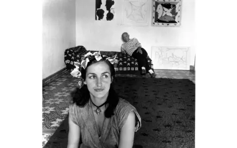 Françoise Gilot joven, con una corona de hojas, mira pensativa mientras Picasso descansa en el fondo, rodeados de sus obras.