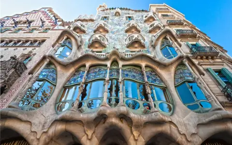 Fachada de edificio **art nouveau** con balcones curvos y vidrieras de colores, reflejando el cielo azul y árboles.