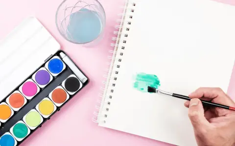 Una mano aplica pintura en acuarela verde en un cuaderno. A su lado, una caja de acuarelas y un vaso con agua azul.