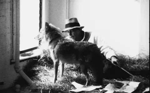 Un hombre con sombrero mira a un lobo junto a una ventana. Me gusta América y América me quiere.