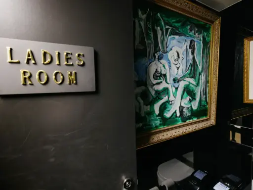 Señal de "Ladies Room" dorada sobre pared oscura, junto a pintura abstracta y retrete.