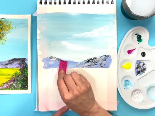 Creando dibujos de paisajes fáciles. Una mano aplica pintura a un lienzo con montañas y cielo azul.