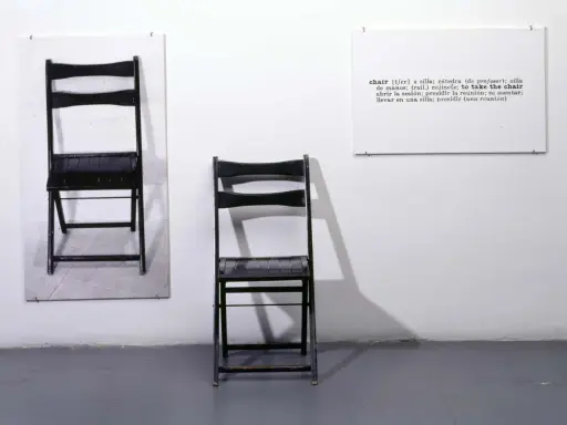 Una y tres sillas. Dos sillas negras plegables, una colgada en la pared y otra de pie, junto a un cartel con la definición de "chair".