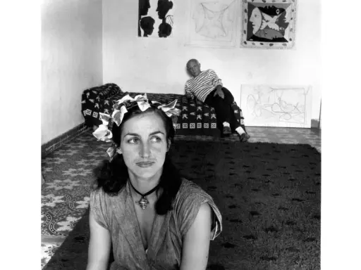Françoise Gilot joven, con una corona de hojas, mira pensativa mientras Picasso descansa en el fondo, rodeados de sus obras.