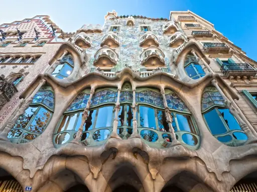Fachada de edificio **art nouveau** con balcones curvos y vidrieras de colores, reflejando el cielo azul y árboles.