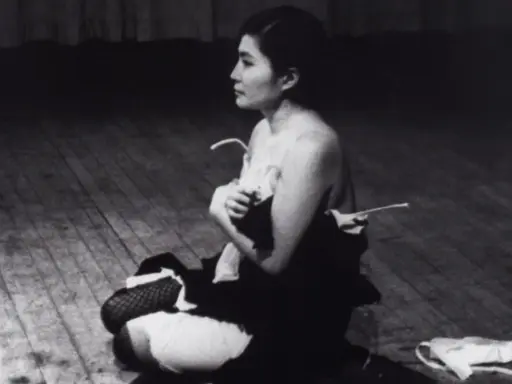 Yoko Ono, en el escenario, sentada en el suelo, con ropa desprendida. Una escena icónica de Yoko Ono Cut Piece.