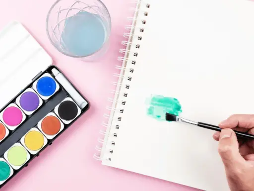 Una mano aplica pintura en acuarela verde en un cuaderno. A su lado, una caja de acuarelas y un vaso con agua azul.