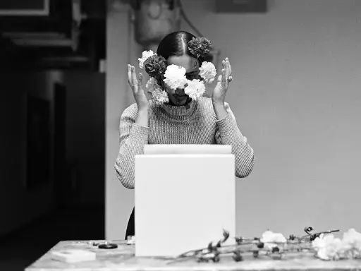 Una mujer con flores en el rostro, evocando las **obras de Ana Mendieta**. Sus manos se alzan, rodeando su cabeza adornada con pétalos blancos y oscuros.