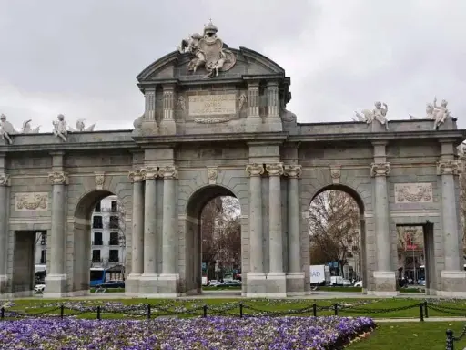 La majestuosa Puerta de Alcalá se alza imponente, con sus arcos y esculturas, rodeada de un jardín de flores moradas.