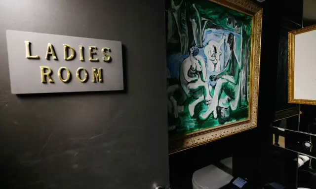 Señal de "Ladies Room" dorada sobre pared oscura, junto a pintura abstracta y retrete.