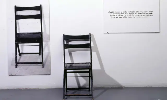 Una y tres sillas. Dos sillas negras plegables, una colgada en la pared y otra de pie, junto a un cartel con la definición de "chair".