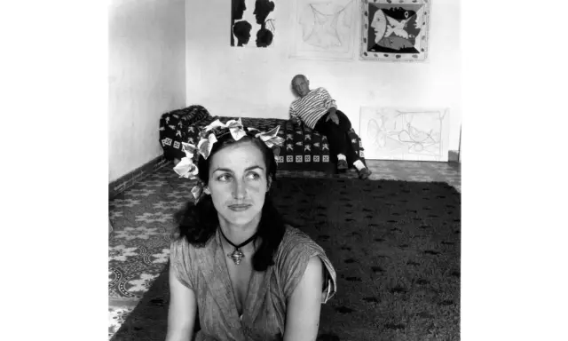 Françoise Gilot joven, con una corona de hojas, mira pensativa mientras Picasso descansa en el fondo, rodeados de sus obras.