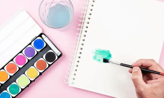 Una mano aplica pintura en acuarela verde en un cuaderno. A su lado, una caja de acuarelas y un vaso con agua azul.