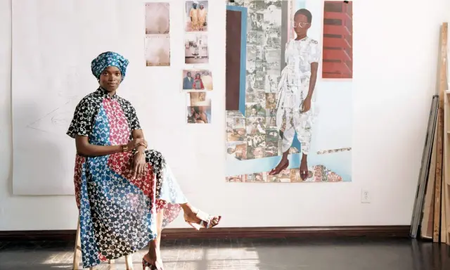 Njideka Akunyili Crosby, sentada en un taburete, luce un vestido vibrante y un turbante. Detrás, obras de arte y fotos familiares adornan la pared.