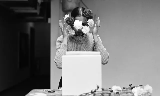 Una mujer con flores en el rostro, evocando las **obras de Ana Mendieta**. Sus manos se alzan, rodeando su cabeza adornada con pétalos blancos y oscuros.