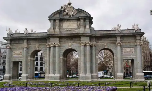 La majestuosa Puerta de Alcalá se alza imponente, con sus arcos y esculturas, rodeada de un jardín de flores moradas.
