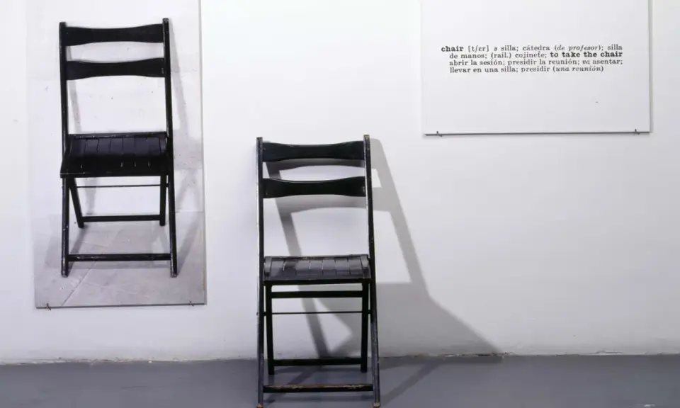 Una y tres sillas. Dos sillas negras plegables, una colgada en la pared y otra de pie, junto a un cartel con la definición de "chair".