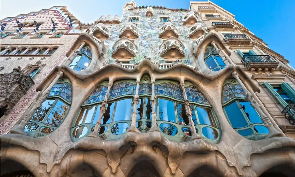 Fachada de edificio **art nouveau** con balcones curvos y vidrieras de colores, reflejando el cielo azul y árboles.
