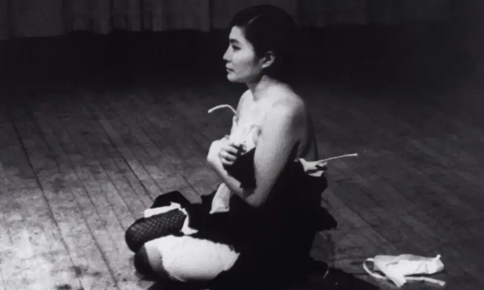Yoko Ono, en el escenario, sentada en el suelo, con ropa desprendida. Una escena icónica de Yoko Ono Cut Piece.