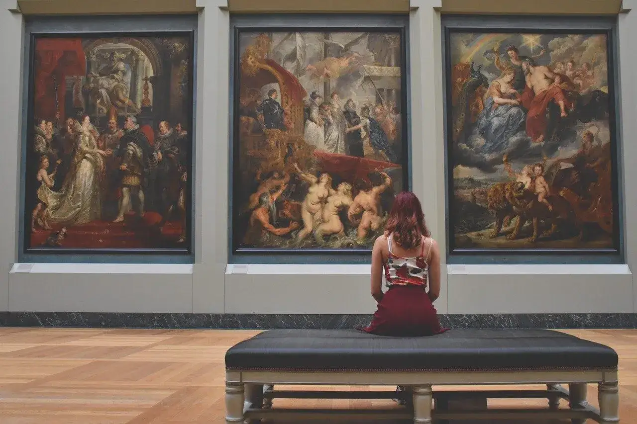 Mujer sentada frente a tres cuadros de museos famosos, admirando obras maestras de Rubens.
