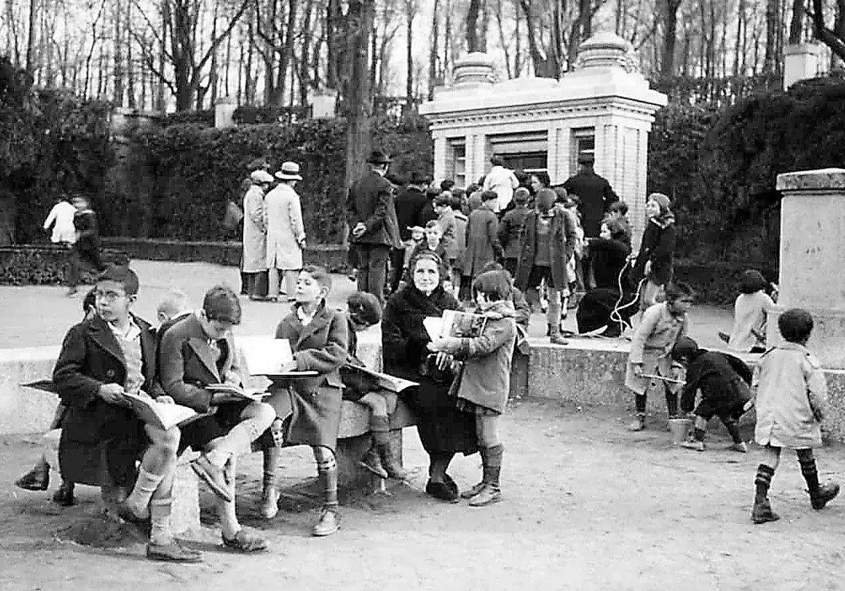 Ni&ntilde;os de 8 a 10 a&ntilde;os disfrutan de la lectura al aire libre en un parque, algunos sentados en un banco, otros de pie, rodeados de &aacute;rboles y un edificio.